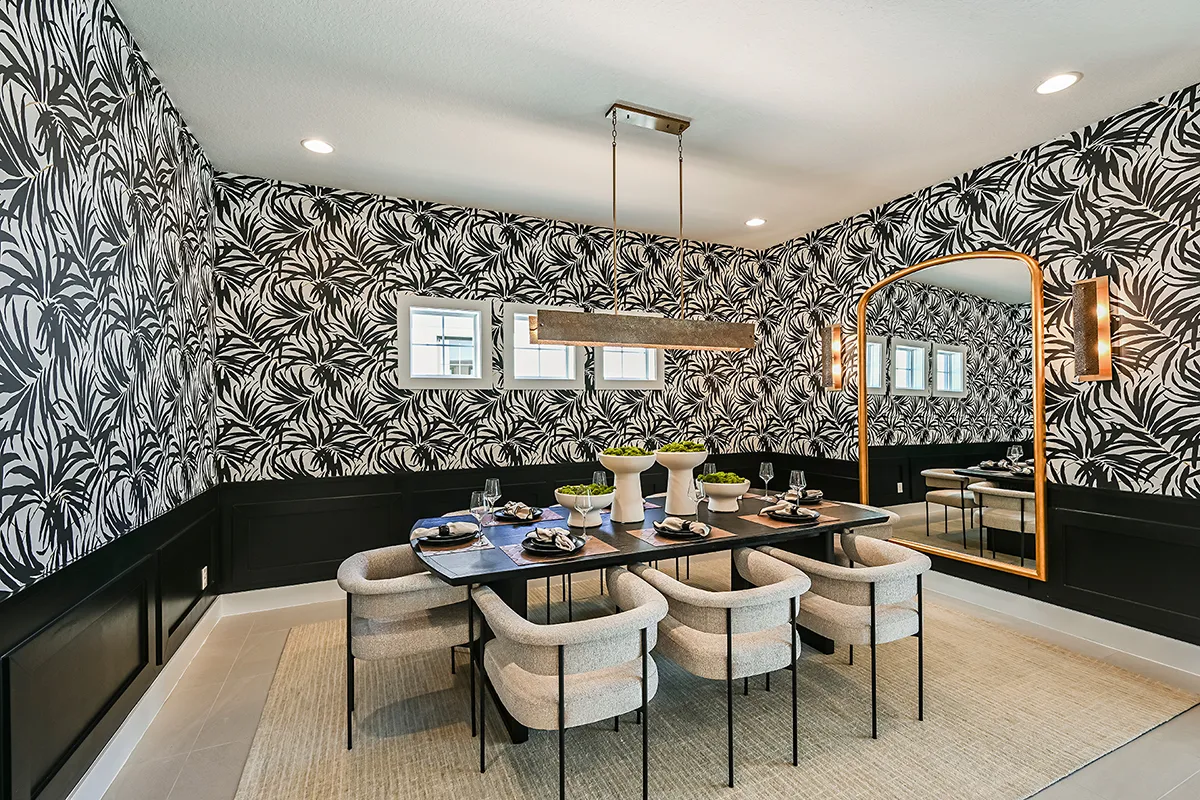 Modern dining room with bold black-and-white leafy wallpaper, a large rectangular table set for eight, cushioned chairs, a gold-framed mirror, and contemporary lighting fixtures, creating an elegant, stylish atmosphere.