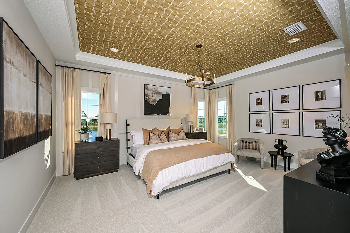 Modern bedroom with a gold-patterned ceiling, large bed with neutral bedding, two nightstands, abstract wall art, a small seating area, and large windows letting in natural light.