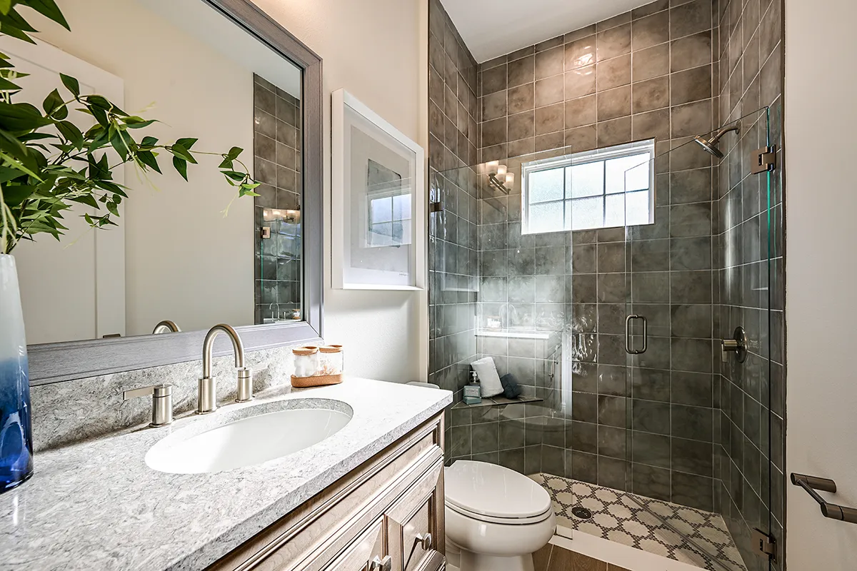 Modern bathroom with a marble-topped vanity, oval sink, silver faucet, potted plant, and a glass-enclosed shower with gray tiles and a window letting in natural light. Toilet and decorative jars are also visible.