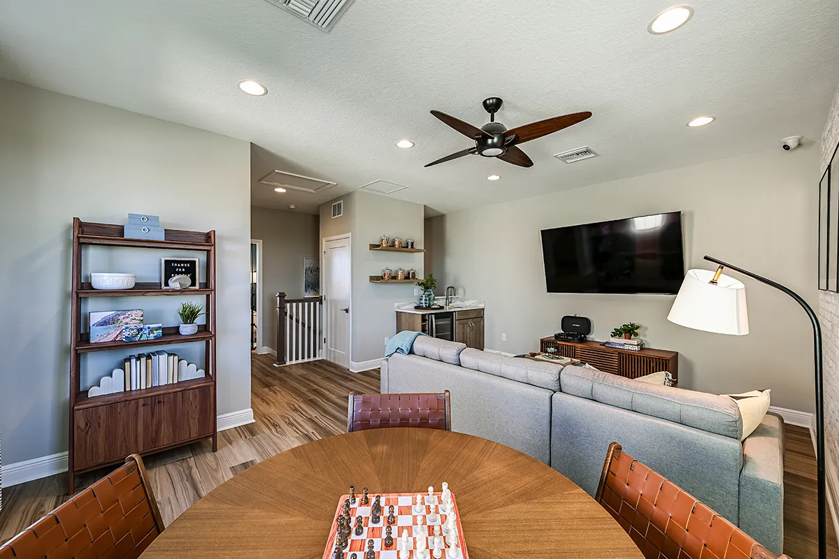Modern living room with light walls, a gray sectional sofa, wall-mounted TV, wooden shelves, and a round table with a chess set. Room has wood flooring, ceiling fan, and decorative accents.