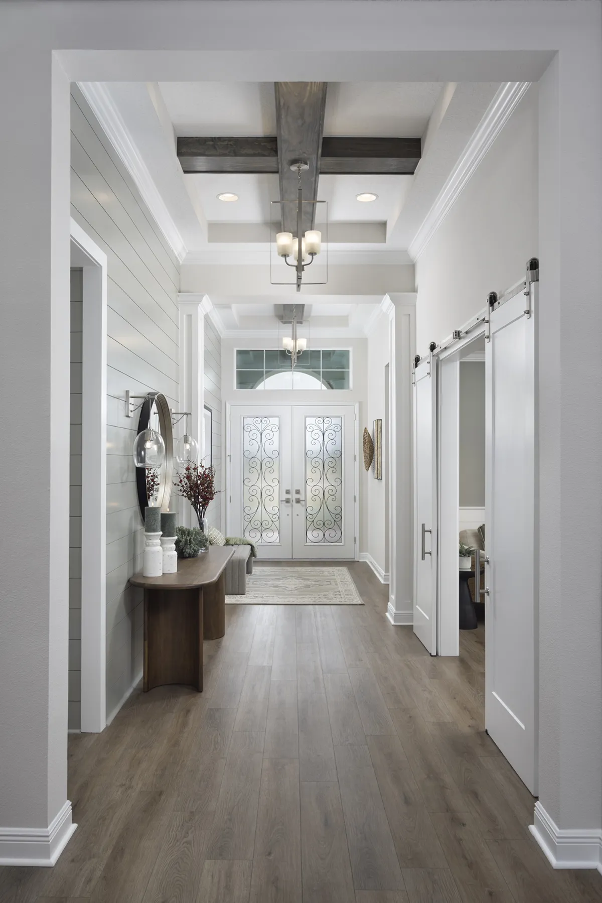 A modern hallway with light wood flooring, white walls, double glass front doors with decorative ironwork, a chandelier, a console table with decor, and white sliding barn doors on the right.