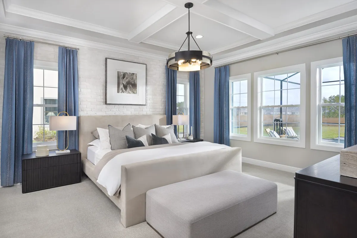 A modern bedroom with a large bed, gray upholstered headboard, blue curtains, white walls, two bedside tables with lamps, a bench at the foot of the bed, and large windows letting in natural light.