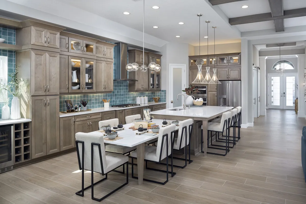 Modern kitchen with wood cabinets, blue tile backsplash, stainless steel appliances, large island with white countertop, six upholstered chairs, pendant lights, wooden floor, and ceiling beams. Bright and spacious design.