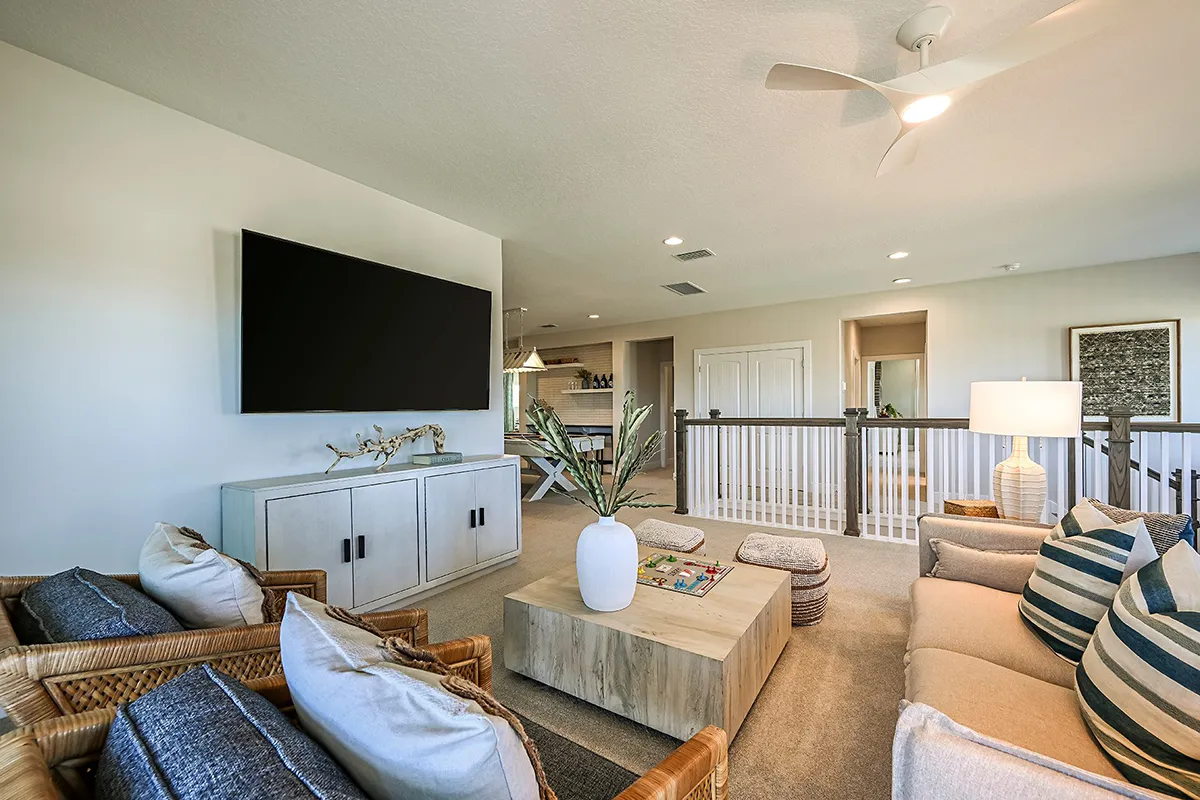 A modern living room with neutral tones features a wall-mounted TV, light wood furniture, cushioned sofas with striped pillows, and a ceiling fan. The space has open railings and connects to other rooms in the background.