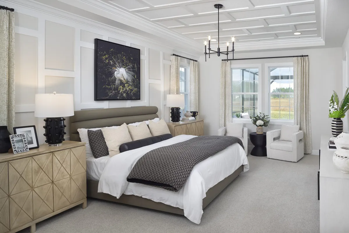 A modern, elegant bedroom with a large bed, neutral bedding, decorative pillows, matching nightstands with lamps, wall art above the bed, armchairs by a window, and a chandelier on a coffered ceiling.
