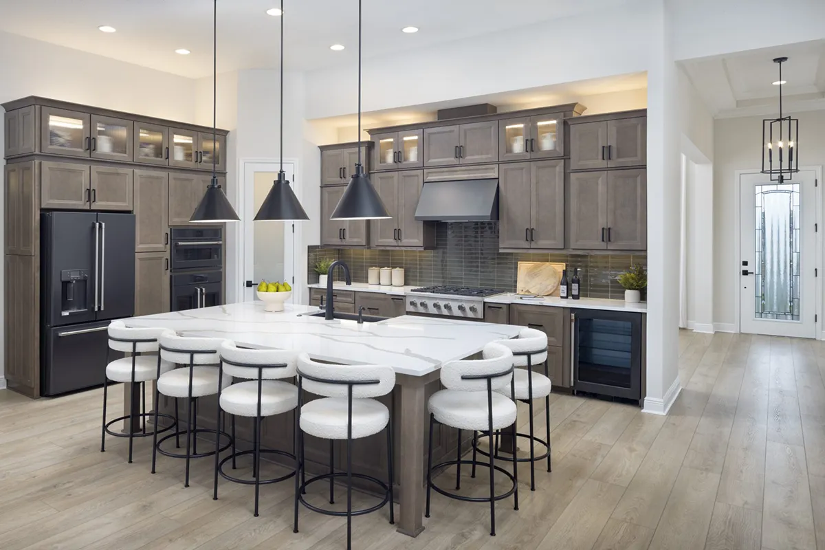 Modern kitchen with gray cabinets, black appliances, a large white island with five round stools, pendant lights above the island, wood flooring, and minimalistic decor. Natural light enters from a door with frosted glass.