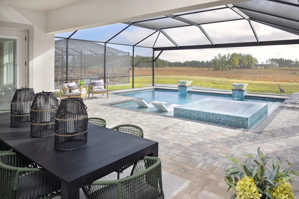 Modern screened-in patio with a dining table and chairs in the foreground; a swimming pool with loungers and water features sits beyond, overlooking an open grassy field and trees in the background.