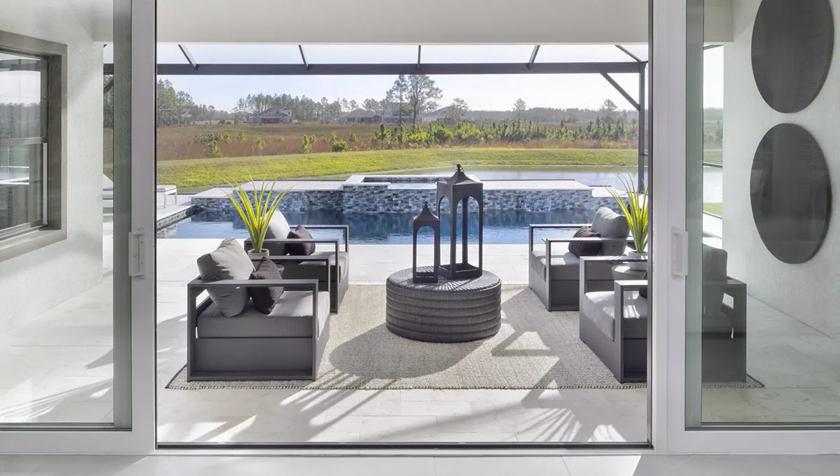 Modern outdoor patio with four gray cushioned chairs around a round woven table, large lantern centerpiece, and rectangular pool in the background, overlooking a grassy landscape under a clear sky.