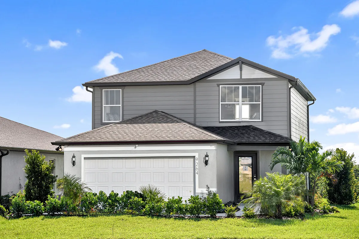 Two-story suburban house with gray siding, white trim, and a double garage. Lush green lawn and shrubs line the front. The sky is clear and blue with a few clouds.