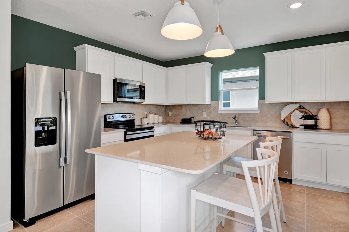 Modern kitchen with white cabinets, stainless steel appliances, an island with two white chairs, pendant lights, and a green accent wall. Countertops are beige, and the floor is tiled. Decorative items sit on the counters.