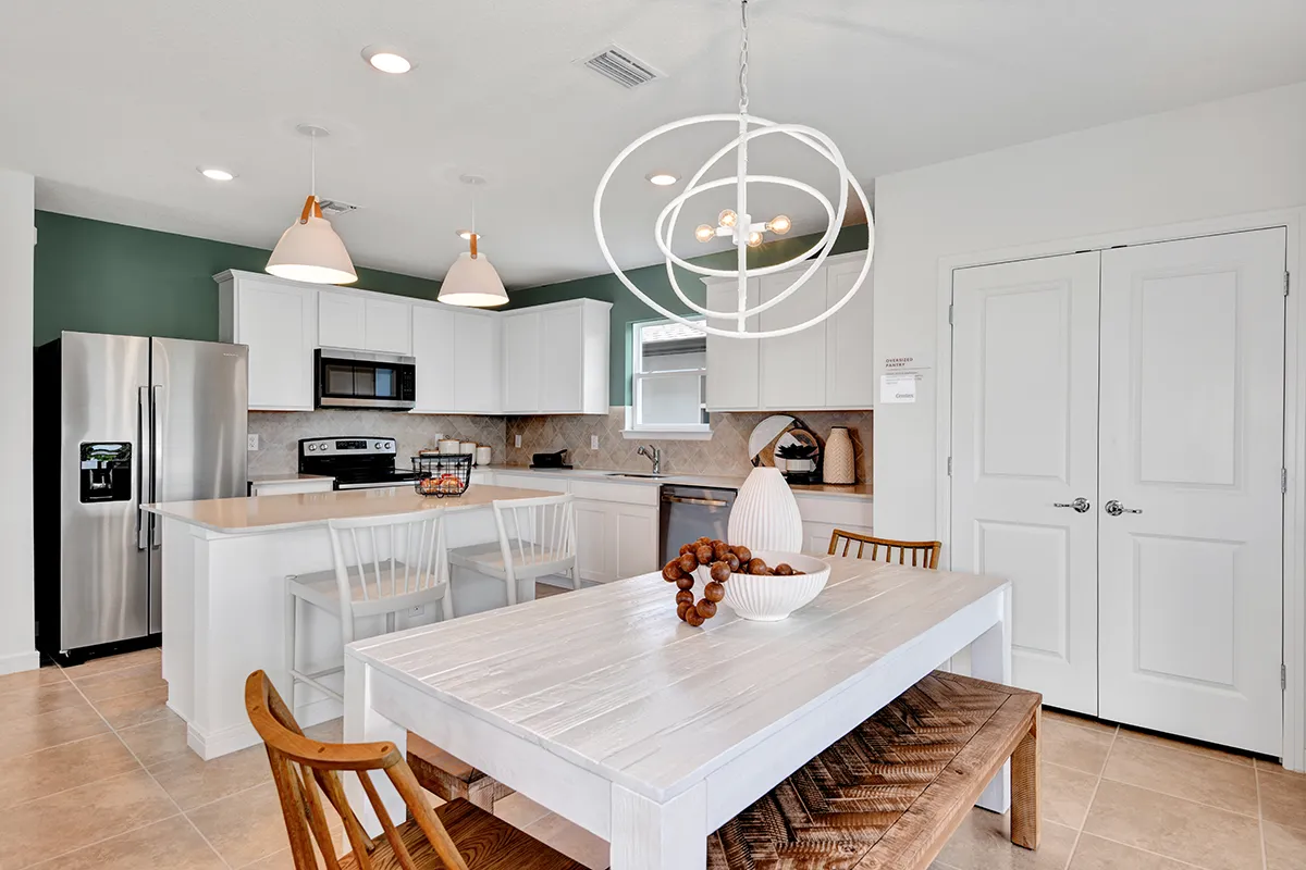 Modern kitchen and dining area with white cabinets, stainless steel appliances, a central island with two chairs, and a white dining table with a bench and chairs under a round, geometric chandelier.