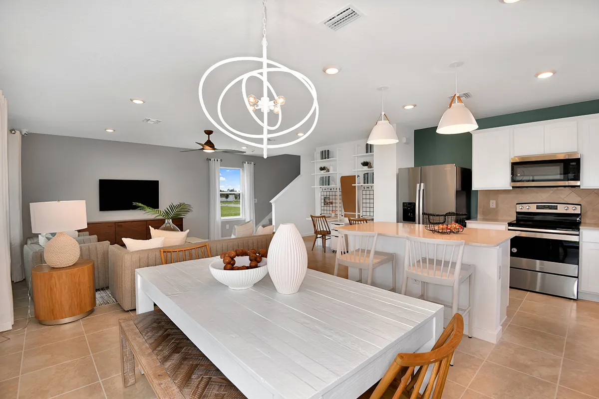 Open-concept kitchen and living area with white cabinets, stainless steel appliances, a breakfast bar with white chairs, a wooden dining table, modern light fixtures, and a cozy sitting area with a TV.
