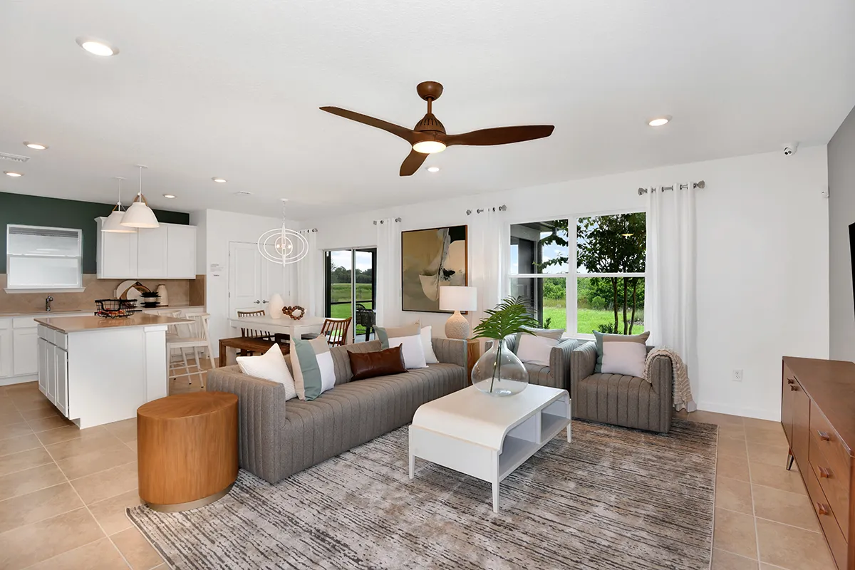 Modern open-concept living room and kitchen with neutral tones, a gray sofa, armchair, large windows with outdoor views, a ceiling fan, minimalist decor, and a dining area with a table and white chairs in the background.