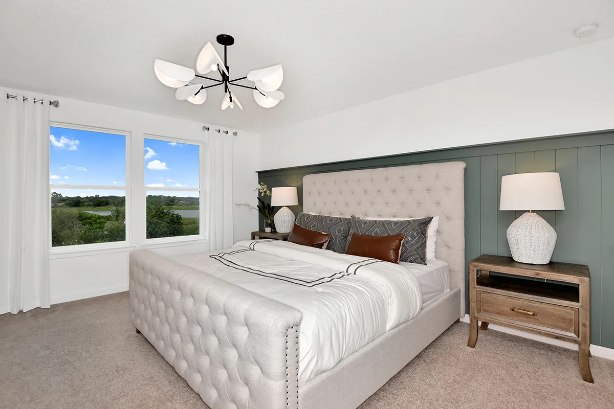 A modern bedroom with a large tufted bed, brown and white bedding, two nightstands with white lamps, green accent wall, beige carpet, white curtains, and a large window with a scenic outdoor view.