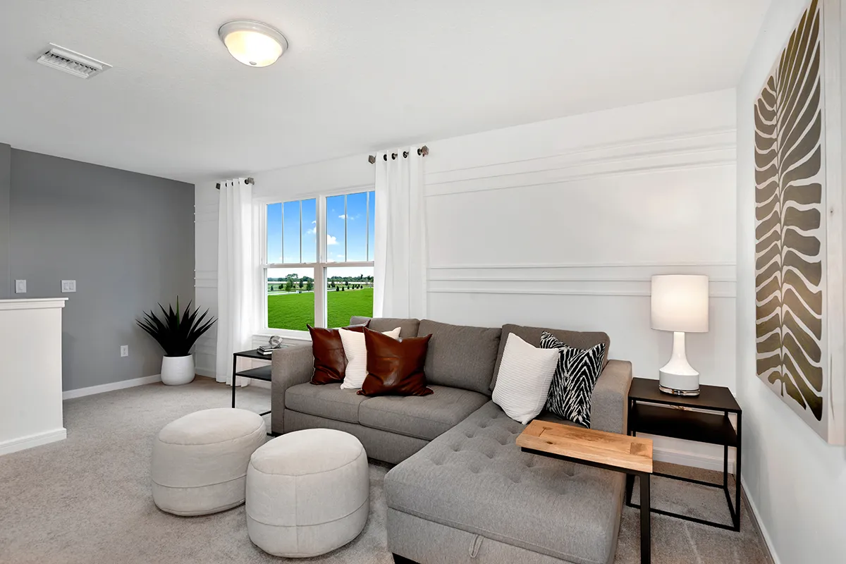 A modern living room with a gray sectional sofa, brown and white pillows, two round ottomans, a wooden side table, a white lamp, potted plants, and a large window with white curtains overlooking a green lawn.