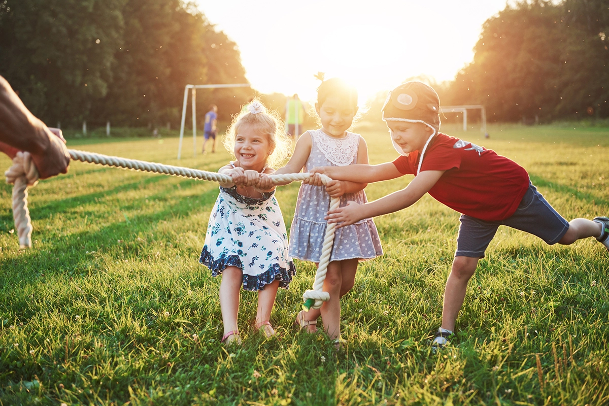 Three children play tug-of-war on a grassy field at sunset, smiling and pulling on a rope together. Soccer goals and other people are visible in the background.