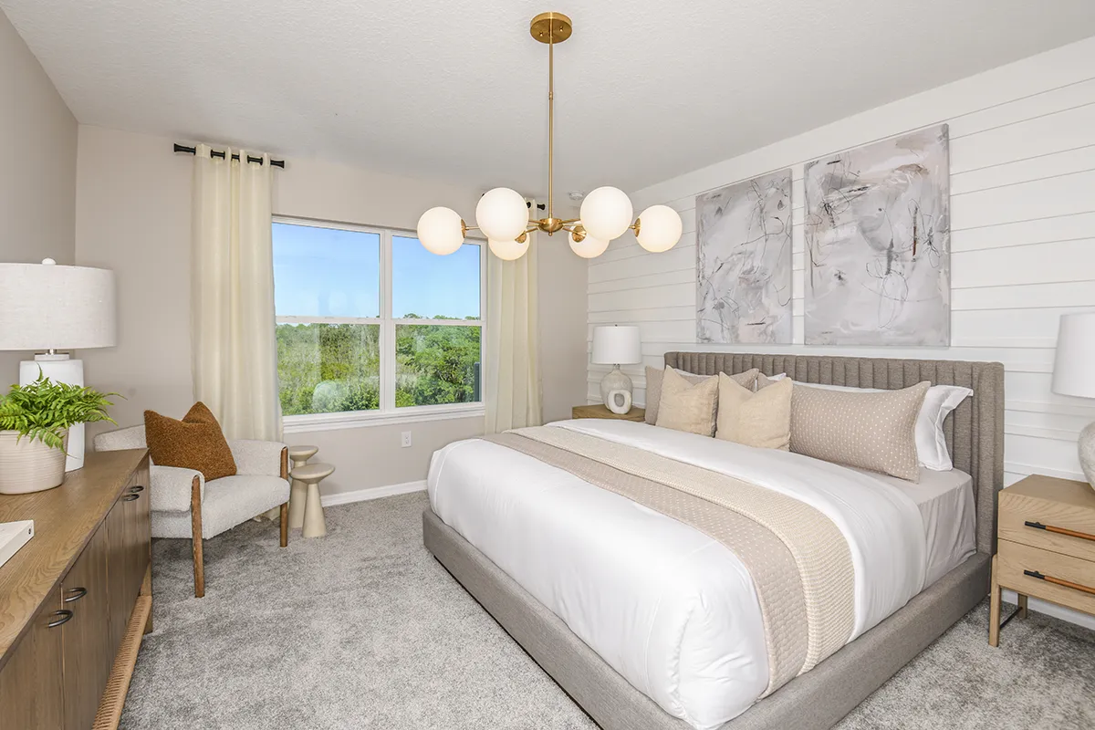 Modern bedroom with a large bed, neutral bedding, two abstract paintings above the headboard, nightstands with lamps, a chair with a pillow, and a window with sheer curtains letting in natural light.