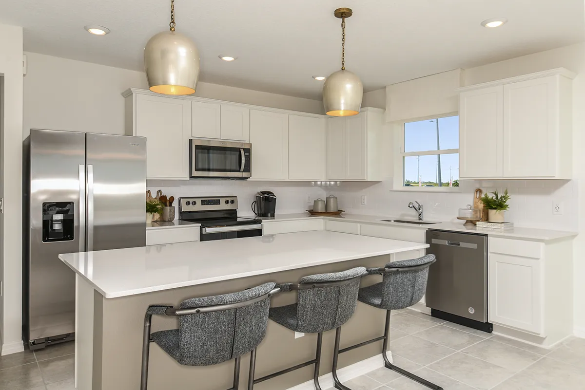 Modern kitchen with white cabinets, stainless steel appliances, a large island with three gray upholstered chairs, pendant lights above, and a window over the sink letting in natural light.