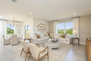 Bright, modern living and dining area with neutral tones, light-colored sofas and chairs, circular coffee table, wall art, large windows with white curtains, and a dining table with upholstered chairs and a gold chandelier.