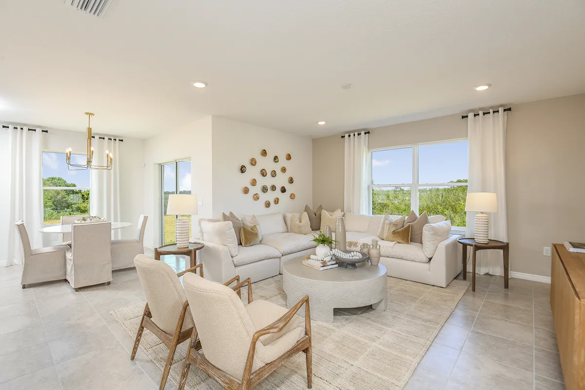 Bright, modern living and dining area with neutral tones, light-colored sofas and chairs, circular coffee table, wall art, large windows with white curtains, and a dining table with upholstered chairs and a gold chandelier.