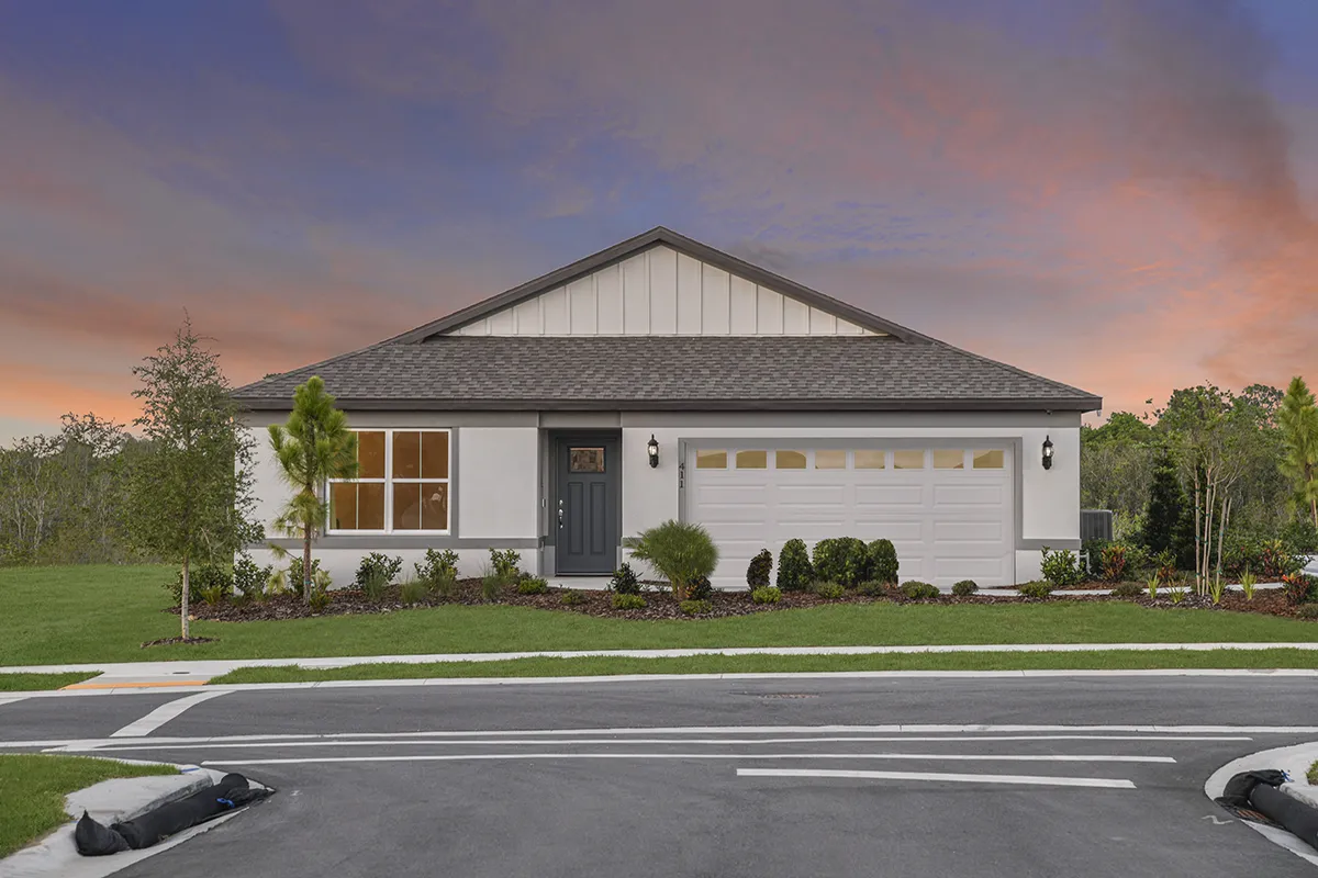 Single-story modern house with a gray roof, white exterior, two-car garage, front lawn, and landscaping, set against a colorful sunset sky. A paved road and sidewalk are visible in the foreground.