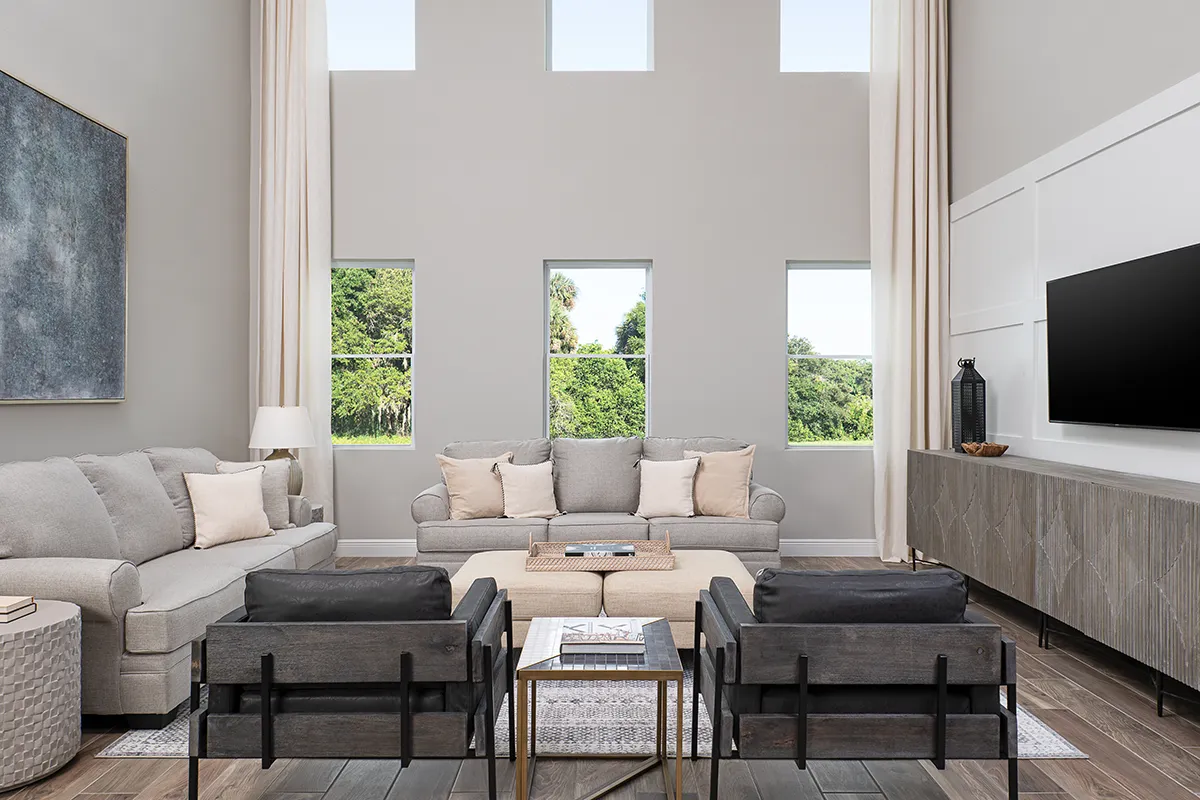 Modern living room with tall ceilings, large windows, neutral-colored sofas, black armchairs, a central ottoman, wall-mounted TV, decorative vases, and natural light streaming in from windows showing greenery outside.