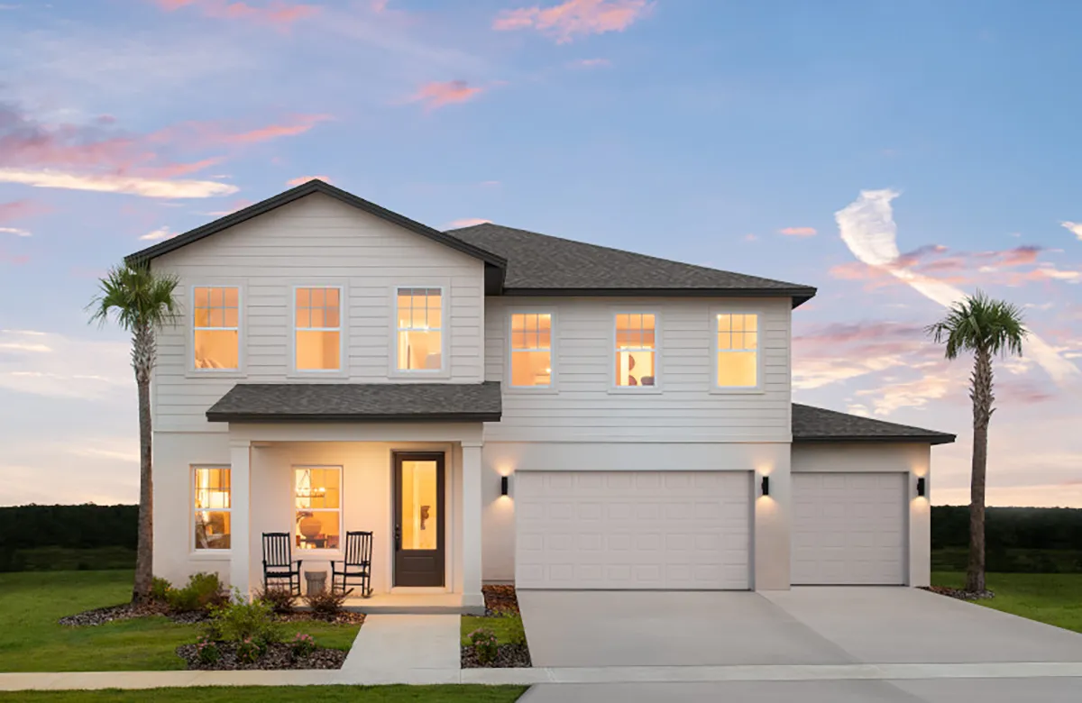 A modern two-story house with white siding, a gray roof, and a three-car garage. Two palm trees stand on either side, and rocking chairs are on the front porch. The sky is blue with pink clouds at sunset.