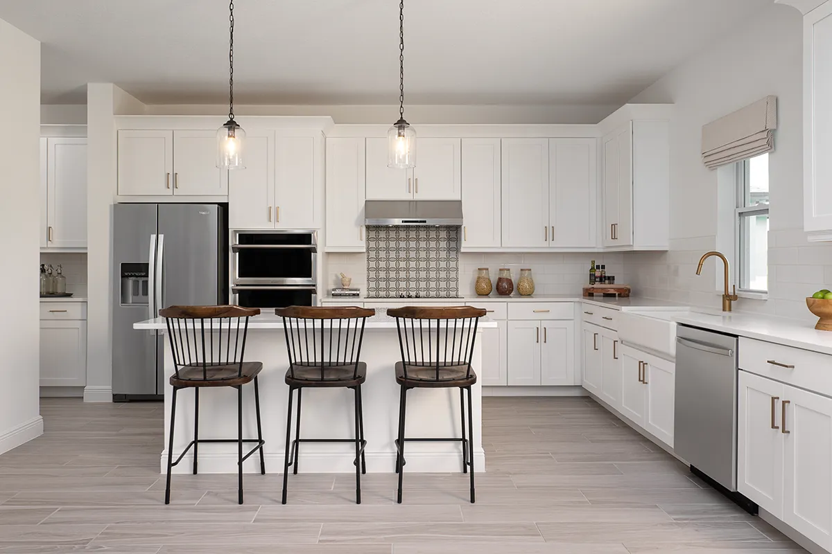 Modern kitchen with white cabinets, stainless steel appliances, a center island with three wooden barstools, pendant lights above, and light gray tile flooring. The space is bright, clean, and uncluttered.