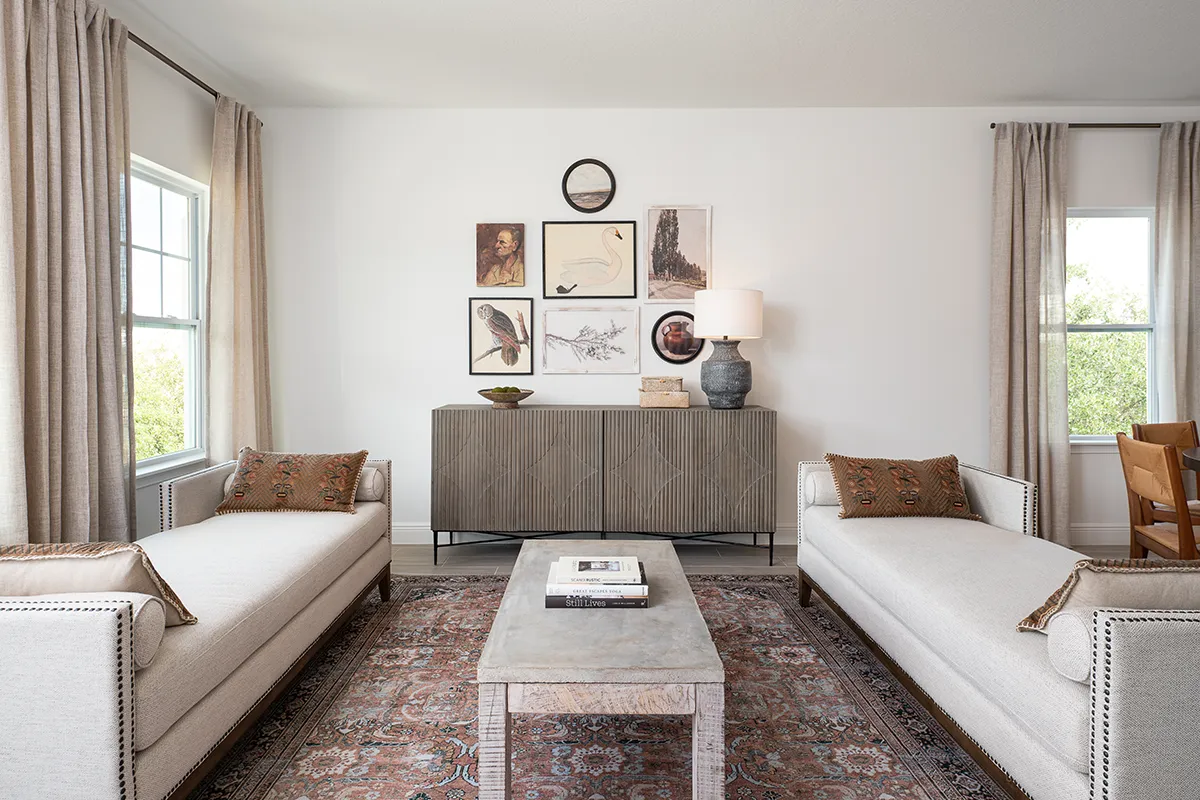 A bright living room with two beige sofas facing each other, a patterned rug, a stone coffee table, a sideboard with a lamp and vase, and framed art on the white wall. Large windows with curtains let in natural light.