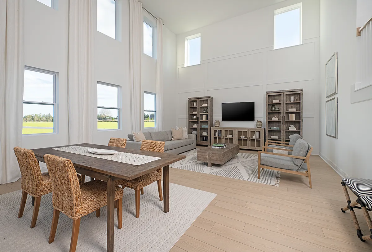 A spacious, modern living and dining area with tall windows, neutral tones, wood floors, a woven dining set, cozy seating, bookshelves, a coffee table, and a wall-mounted TV. Natural light fills the room.