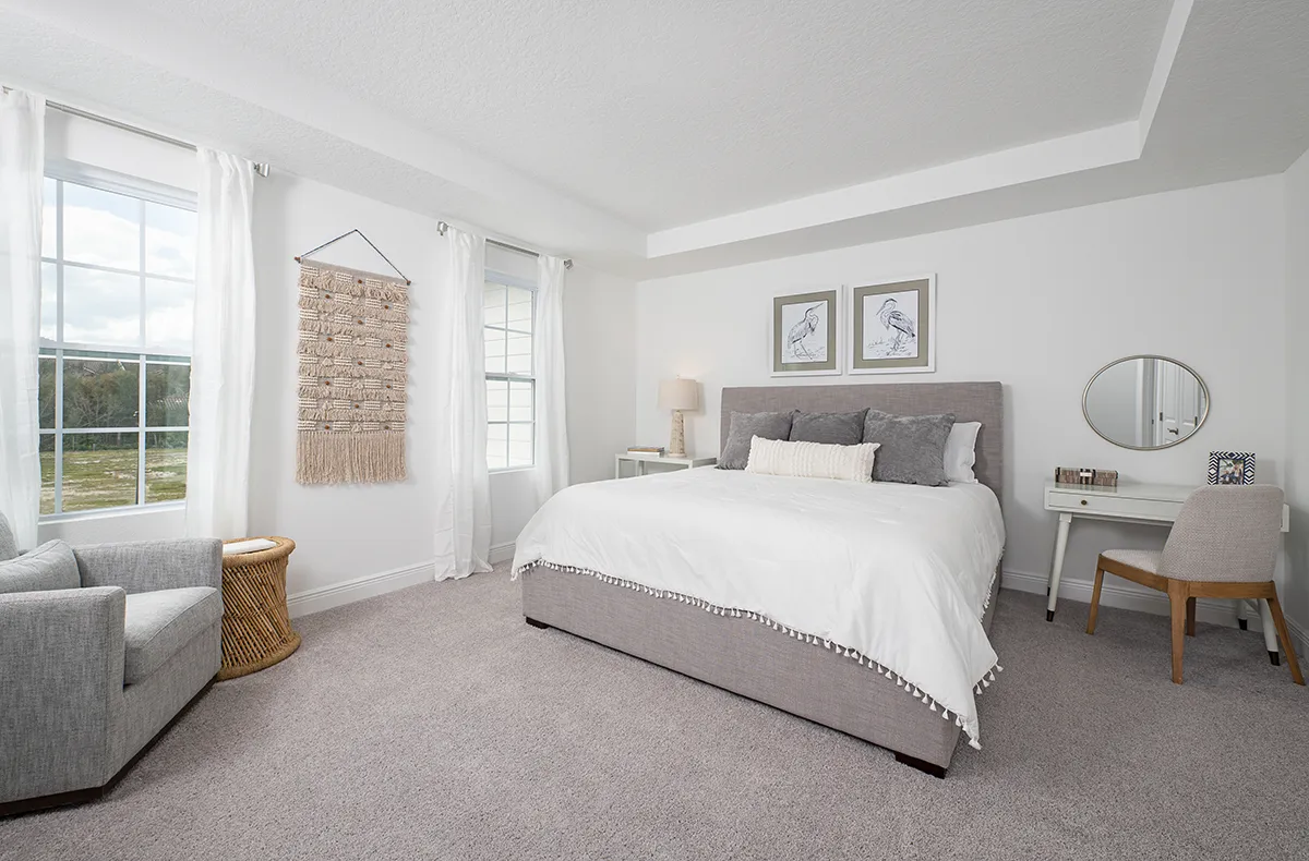 Bright, modern bedroom with large windows, gray upholstered bed with white bedding, a gray armchair, woven side table, macramé wall hanging, small desk with chair, lamp, mirror, and framed bird art on white walls.