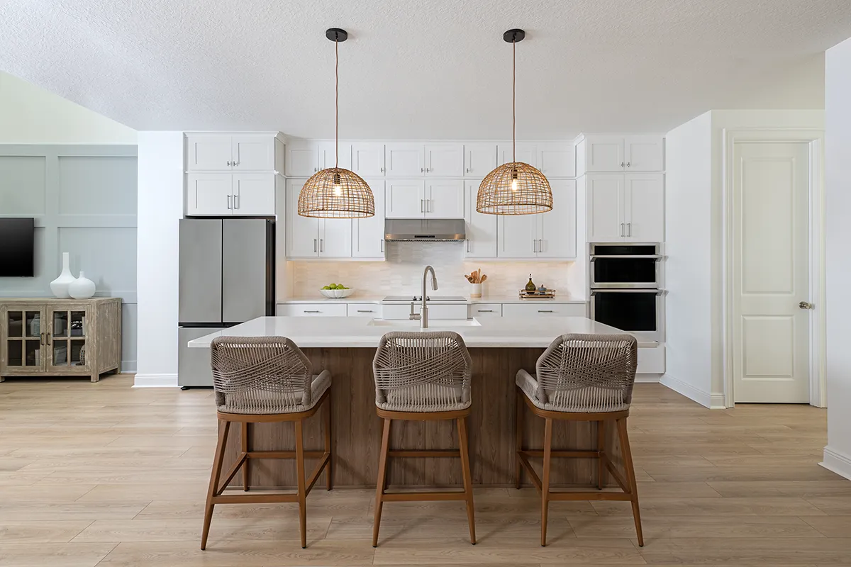 Modern kitchen with white cabinets, stainless steel appliances, and a central island featuring three wicker barstools and two woven pendant lights. The space has wood flooring and a neutral, minimalist design.