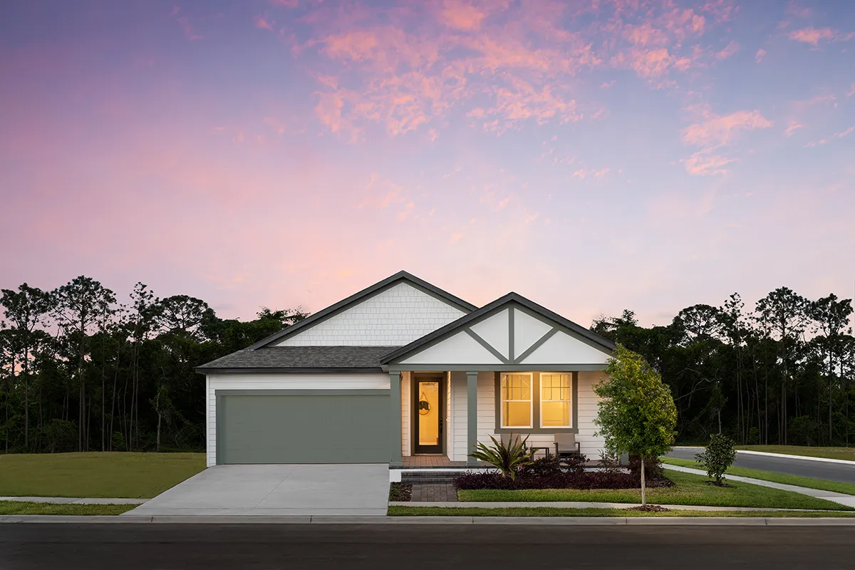 A modern single-story house with a two-car garage, well-kept lawn, illuminated front porch, and decorative landscaping, set against a sunset sky with pink clouds and trees in the background.