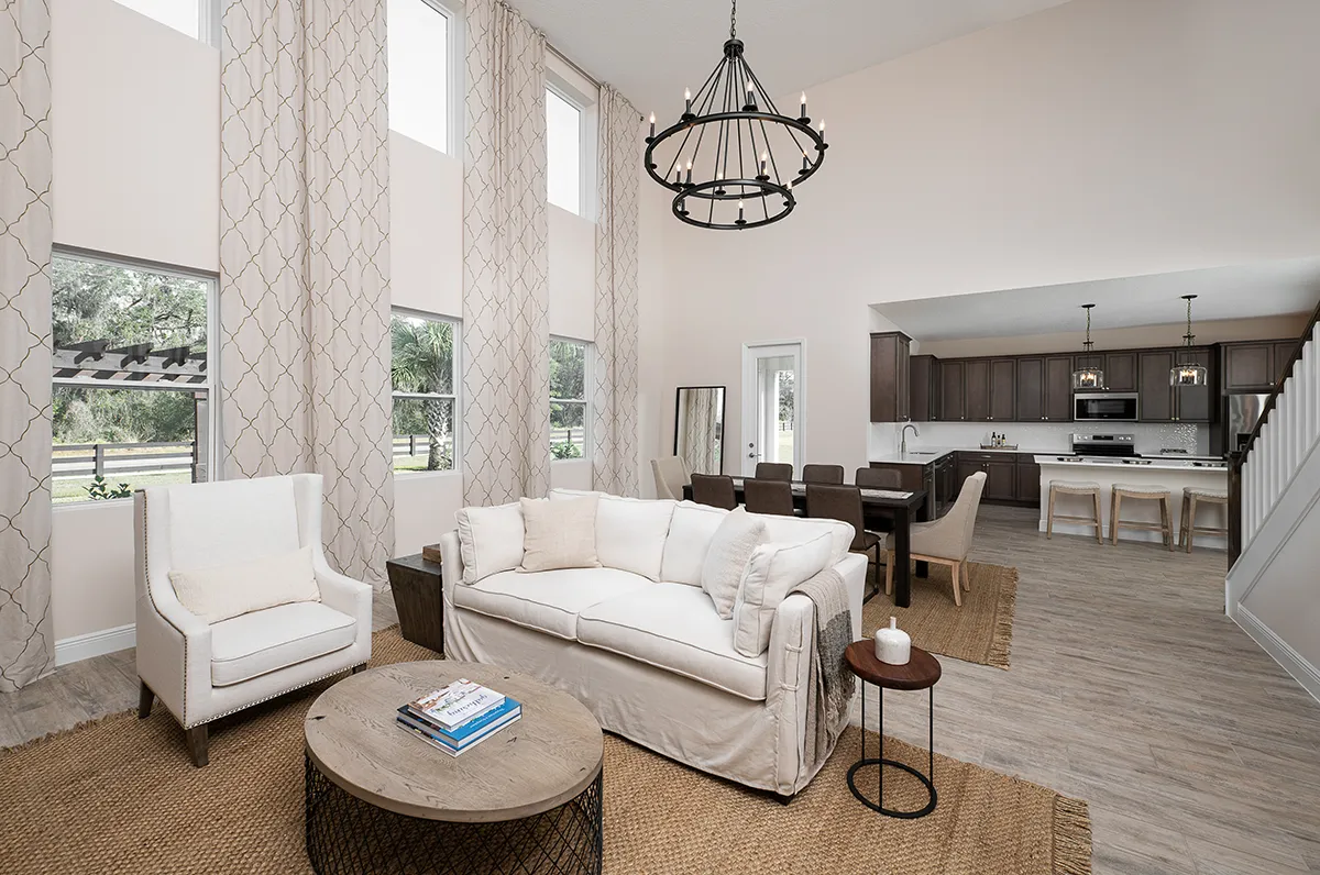 A modern, open-concept living and dining area with tall windows, neutral décor, a white sofa, armchair, wood coffee table, dining set, and a kitchen in the background with dark cabinets and stainless steel appliances.