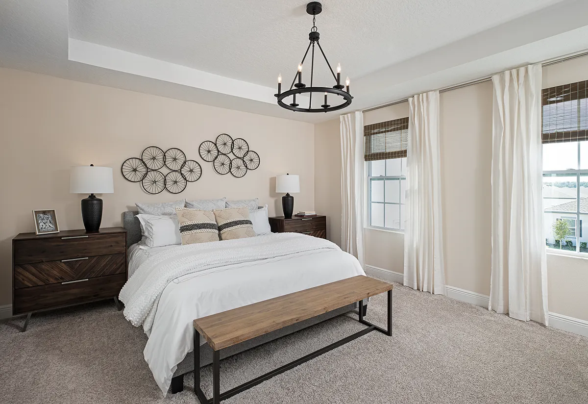 A bright, modern bedroom with a neatly made bed, wooden bench, two dark nightstands with lamps, decorative wall art, light carpet, large windows with curtains, and a black chandelier overhead.