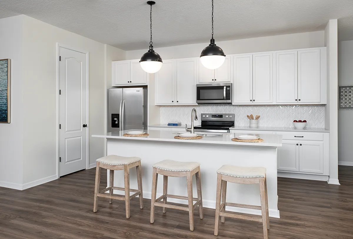 Modern white kitchen with stainless steel appliances, island with sink, three wooden stools, two pendant lights above, and wood flooring. Cabinets and drawers line the back wall, with minimal decor and a clean, bright look.