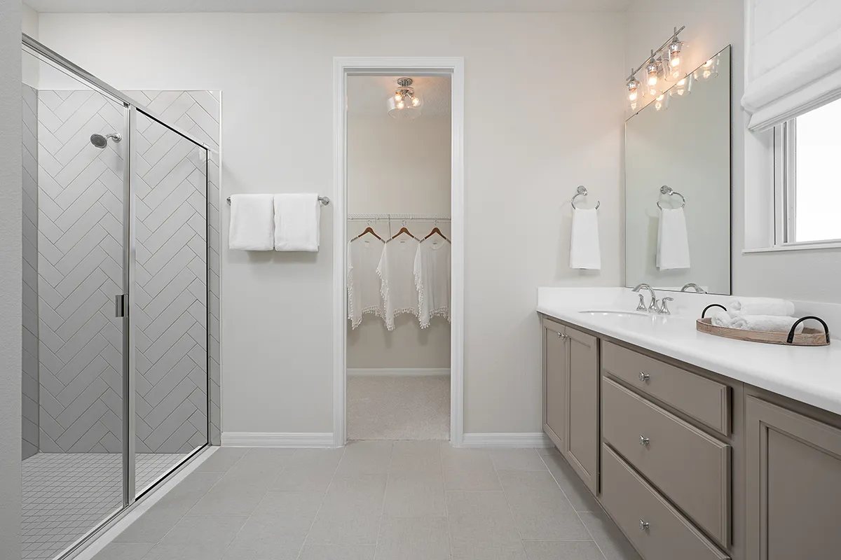 Modern bathroom with a glass shower on the left, double sink vanity with gray cabinets on the right, white towels, and an open door leading to a walk-in closet with hanging white shirts.