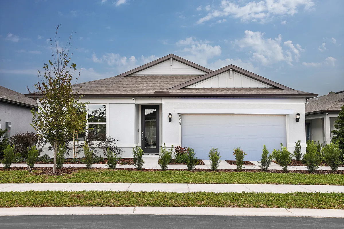 A single-story modern suburban house with white exterior walls, gray roof, two-car garage, small front yard with shrubs, and a young tree, set against a partly cloudy sky.