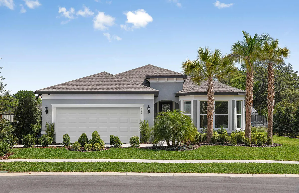 Single-story suburban house with a gray exterior, white trim, and a two-car garage. The front yard features manicured lawns, shrubs, and several palm trees under a blue sky with scattered clouds.