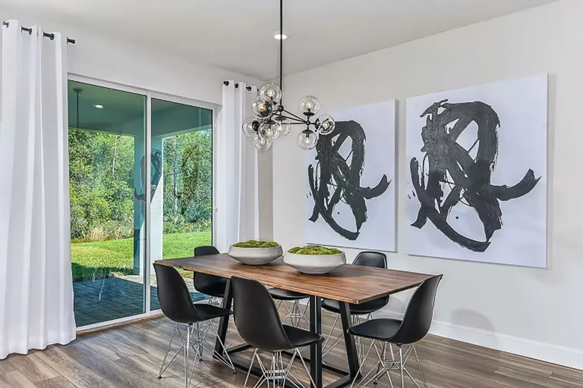 Modern dining room with a wooden table, six black chairs, two abstract black-and-white paintings on the wall, contemporary chandelier, large window with white curtains, and view of greenery outside.