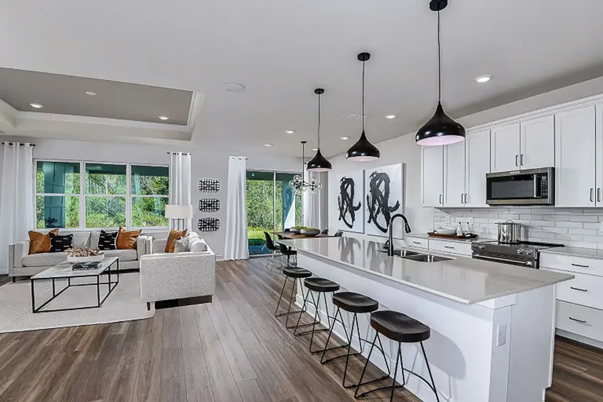 Modern open-concept kitchen and living room with white cabinets, pendant lights over a long island with four stools, stainless steel appliances, and a cozy sitting area with sofas and large windows letting in natural light.