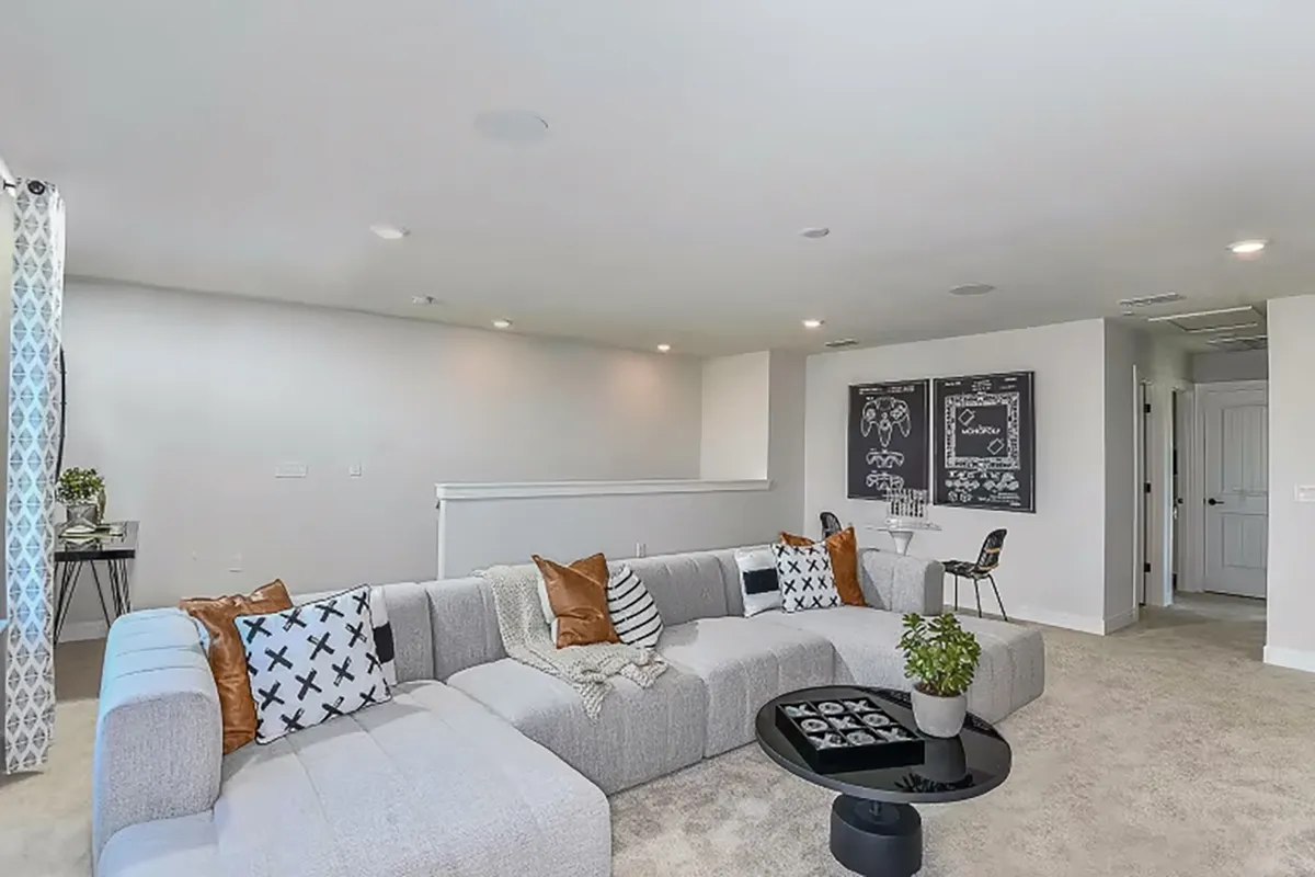 A spacious living room with a large gray sectional sofa, patterned throw pillows, a round black coffee table with a plant, and a workspace with two chalkboard-style art pieces on the wall in the background.