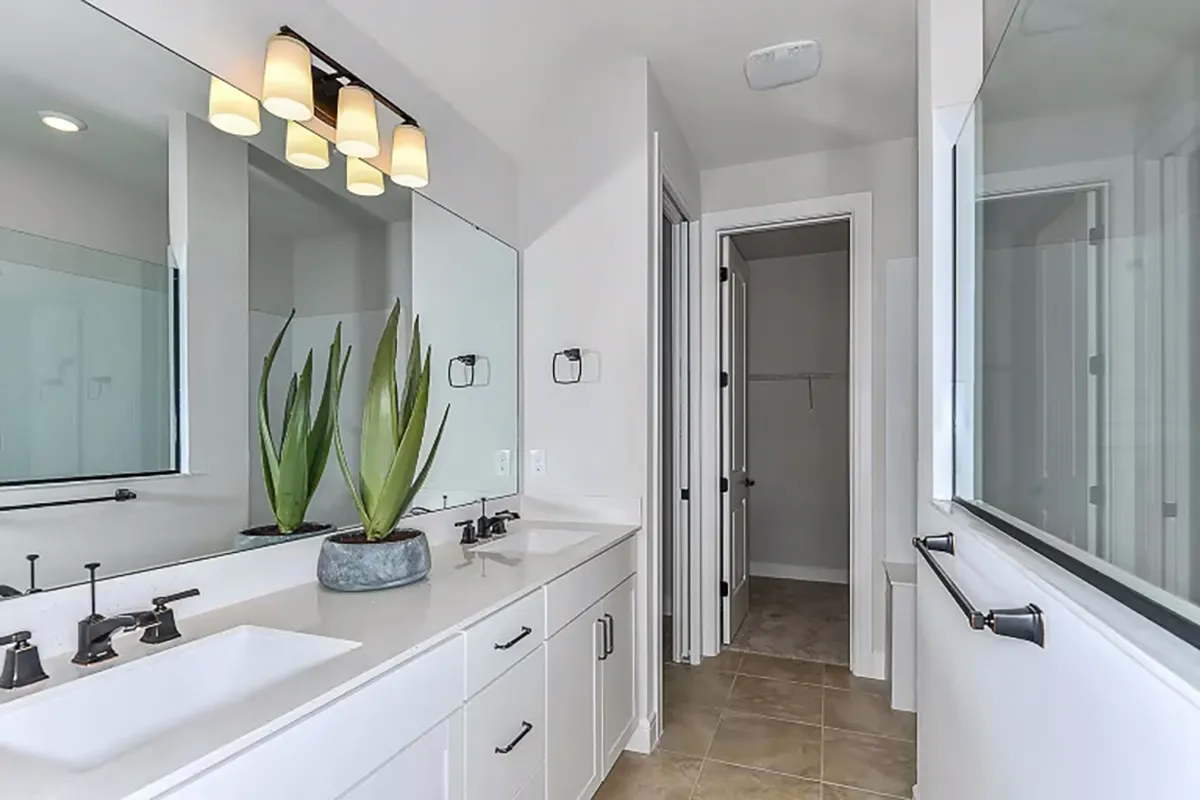 Modern bathroom with double sinks, white cabinets, large mirror, black fixtures, and a potted plant on the counter. Theres a glass shower to the right and an open door leading to a walk-in closet.