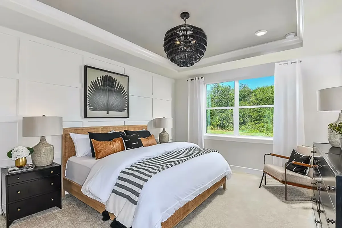 Modern bedroom with a large bed, decorative pillows, two bedside tables with lamps, a black and white feather artwork above the headboard, a window with white curtains, and a black chandelier.
