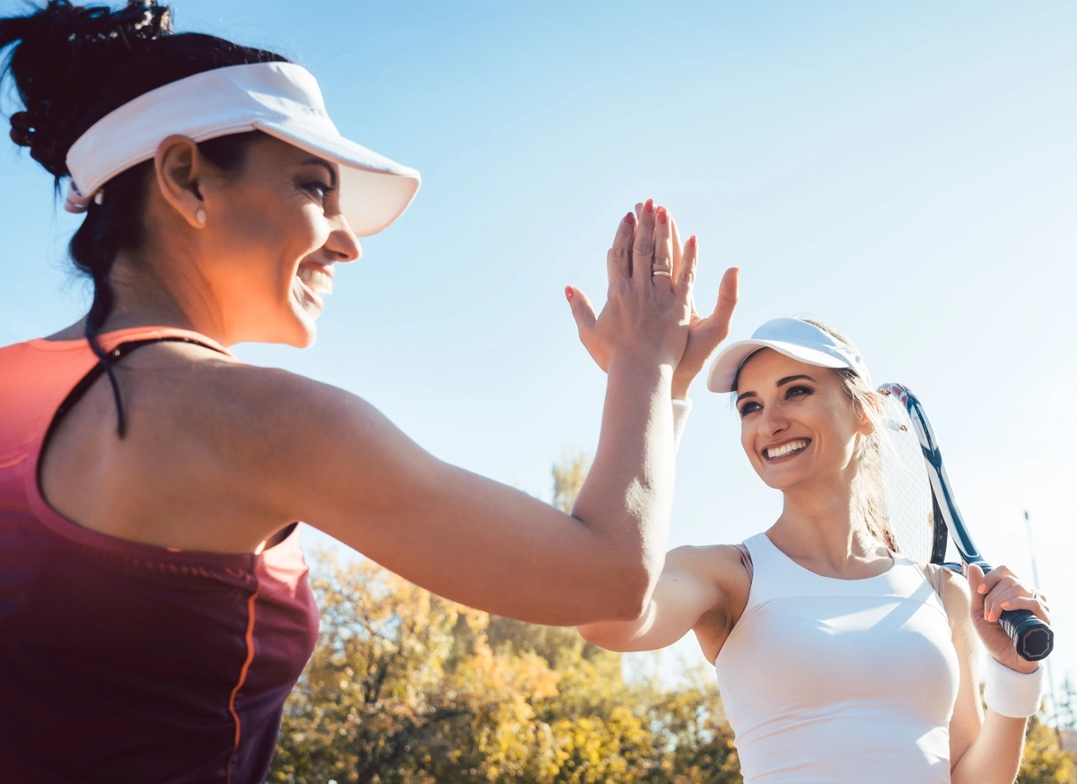 Two women wearing sports visors and athletic clothing smile and high-five each other outdoors on a sunny day. One woman holds a tennis racquet, and trees are visible in the background.