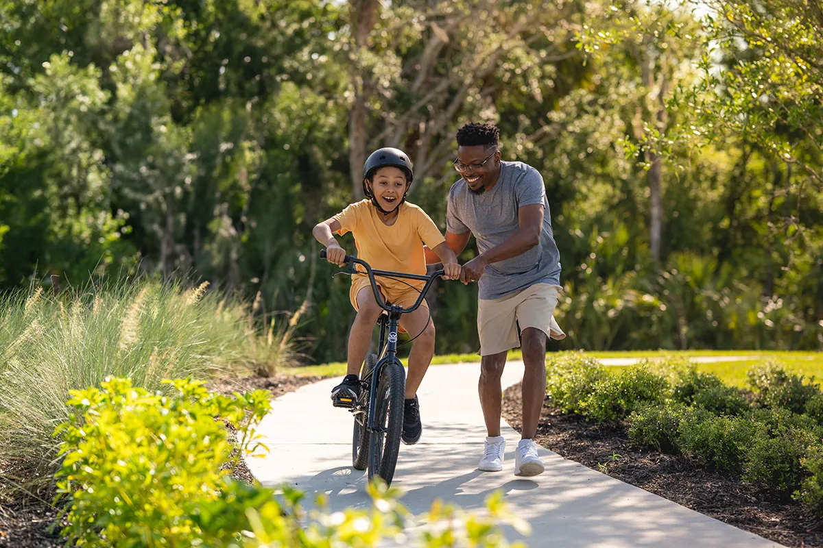 A smiling adult helps a child ride a bicycle on a sunny park path. The child wears a helmet and looks excited, while the adult runs alongside, holding the bike. Green trees and bushes surround the path.