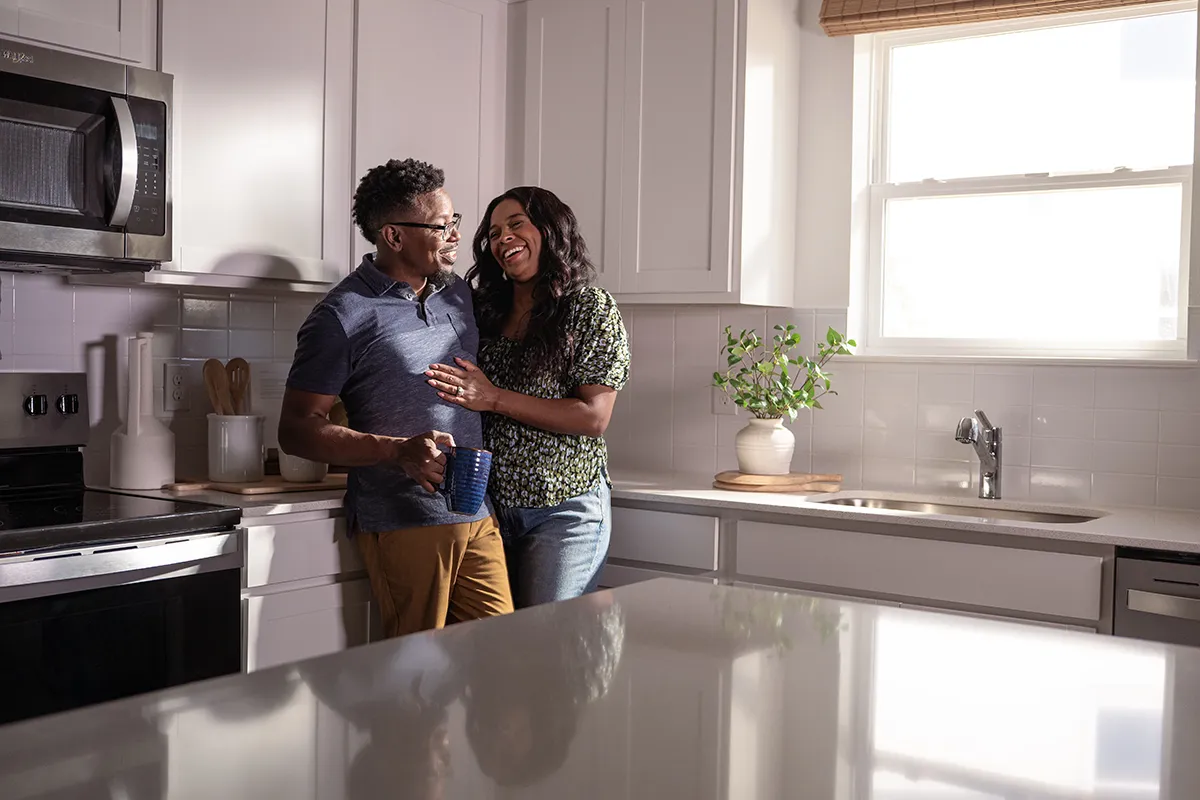 A couple stands together in a bright, modern kitchen, smiling and talking. One person holds a blue mug, and sunlight streams in through a window above a sink with a potted plant nearby.