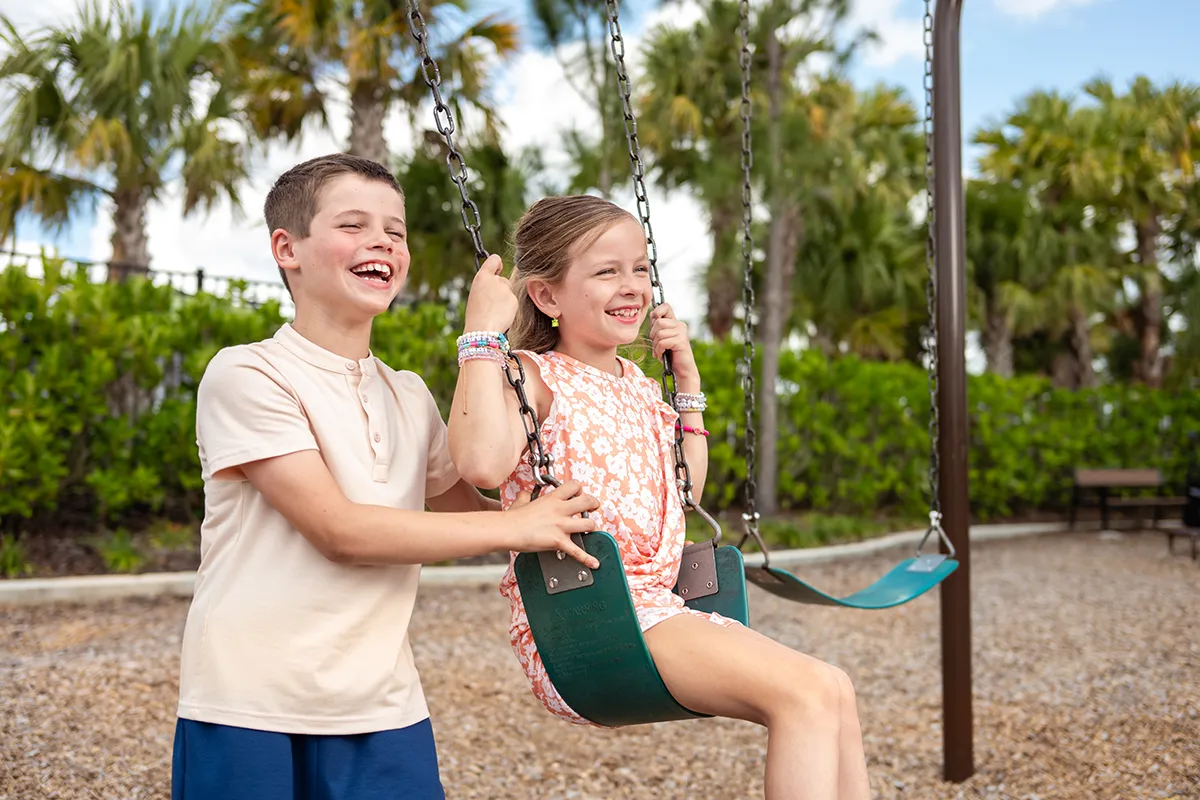 A smiling boy pushes a smiling girl on a swing at a playground, surrounded by green bushes and palm trees on a sunny day.