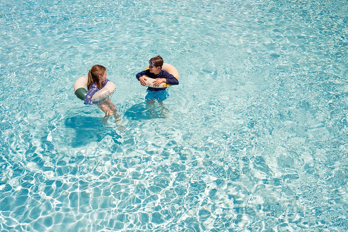 Two children wearing swim shirts and using inflatable rings float and talk together in a clear blue swimming pool, with sunlight creating patterns on the waters surface.