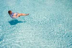 A person in a blue and white swimsuit floats on their back in a clear, sparkling swimming pool, surrounded by bright, rippling water.